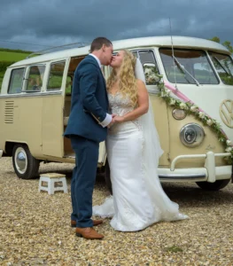 Bride and groom sharing a kiss beside a vintage VW camper van decorated with flowers.