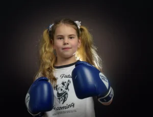 Studio portrait of a young girl wearing boxing gloves, lit dramatically against a dark background.