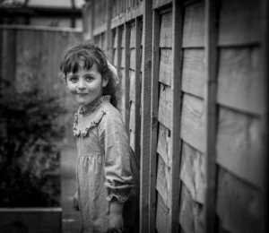 Black & white image of a young girl standing near a garden fence in