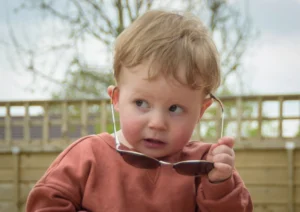 Toddler boy wearing sunglasses with a curious expression, photographed in a natural outdoor setting.