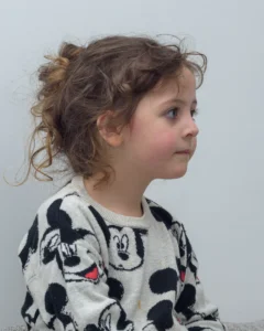 Side-profile portrait of a little girl with tousled hair wearing a Mickey Mouse jumper, taken during a relaxed indoor photoshoot.