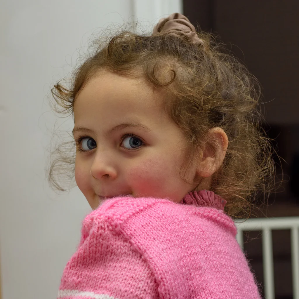 Esme shoulder pose-1 Little girl in a pink knitted jumper glancing back over her shoulder with a playful smile.