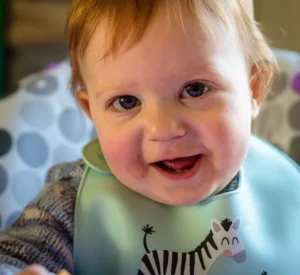 Happy baby boy with a bib smiling at the camera, captured during a natural light baby photography session.