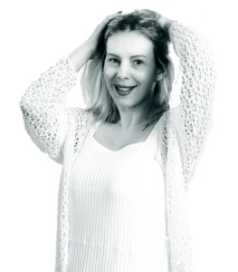 High-key black-and-white studio portrait of a woman smiling with her hands in her hair.