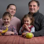 Family seated at a table with fruit, smiling and interacting playfully.
