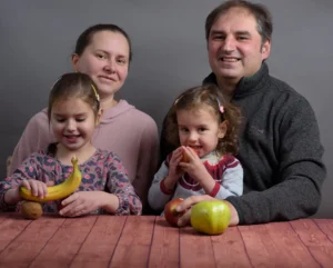 Family seated at a table with fruit, smiling and interacting playfully.