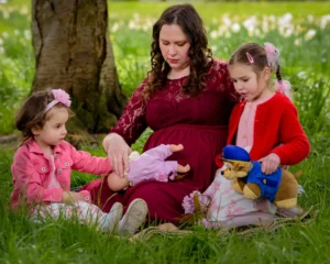 Mother sitting with two young daughters beneath cherry blossom trees, playing with a toy doll