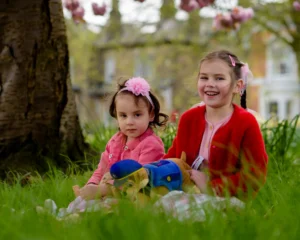 wo young sisters sitting in the grass beneath pink cherry blossoms, one smiling brightly.